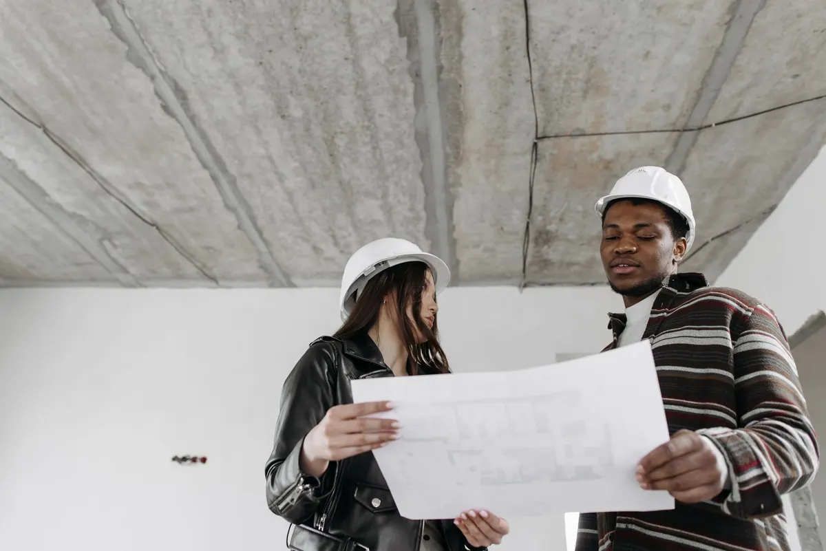 Deux professionnels casqués lisent un plan dans un bâtiment en cours de gros œuvre, lors d'une visite de chantier