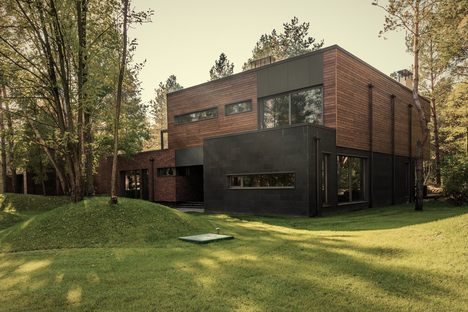 Maison contemporaine à étage en bardage bois et rez-de-chaussée sombre, sur une pelouse bordée de pins, photo d'illustration
