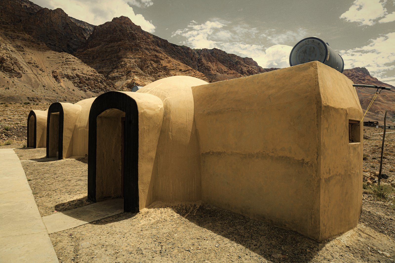 Bâtiment contemporain aux murs en terre enduite et portes cintrées, sur un terrain sec au pied de montagnes, photo d'illustration