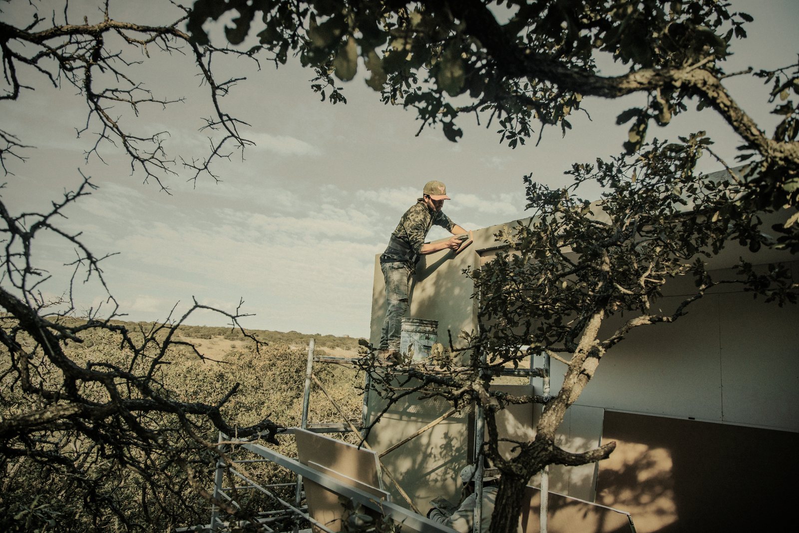 Artisan vu de loin sur un échafaudage, travaillant sur la façade d'une maison entourée d'arbres, photo d'illustration