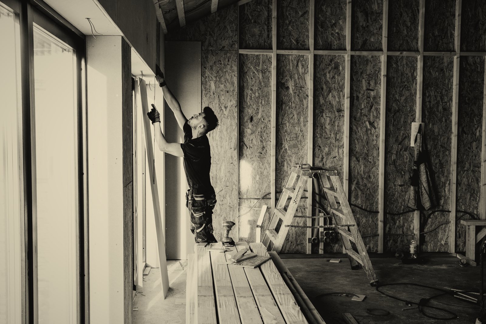 Ouvrier de dos fixant des panneaux sur une ossature bois à l'intérieur d'une maison en chantier, photo d'illustration