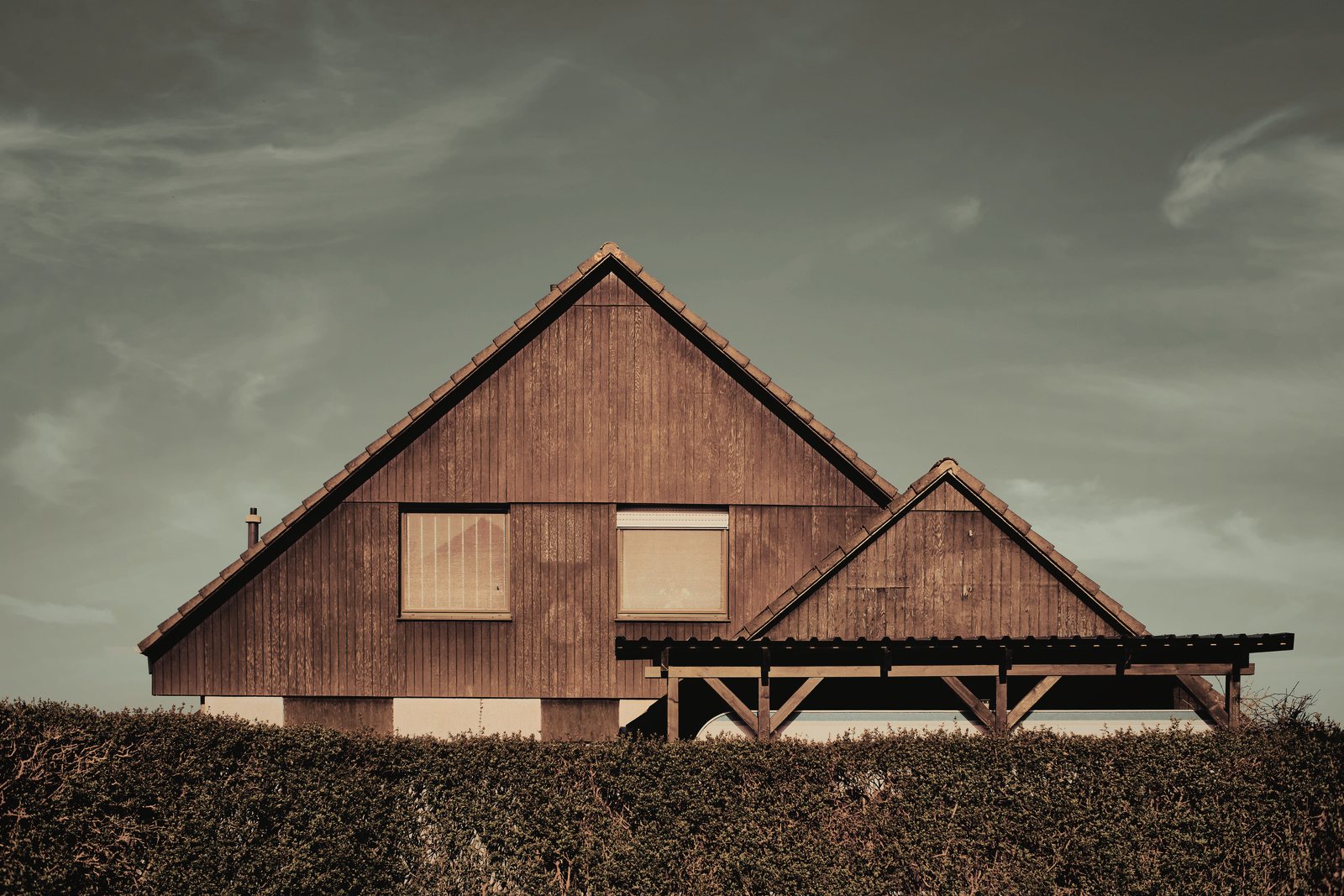 Maison à grands pignons en bardage bois brun derrière une haie, sous un ciel couvert, photo d'illustration