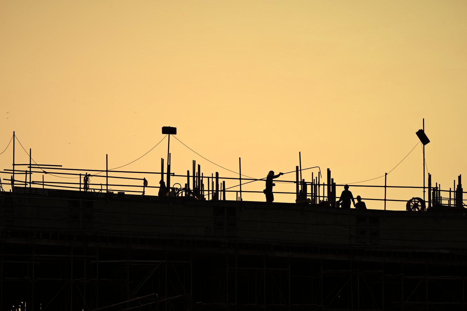 Silhouettes d'ouvriers sur la dalle d'un bâtiment en construction, garde-corps et étais, ciel orangé du soir, photo d'illustration