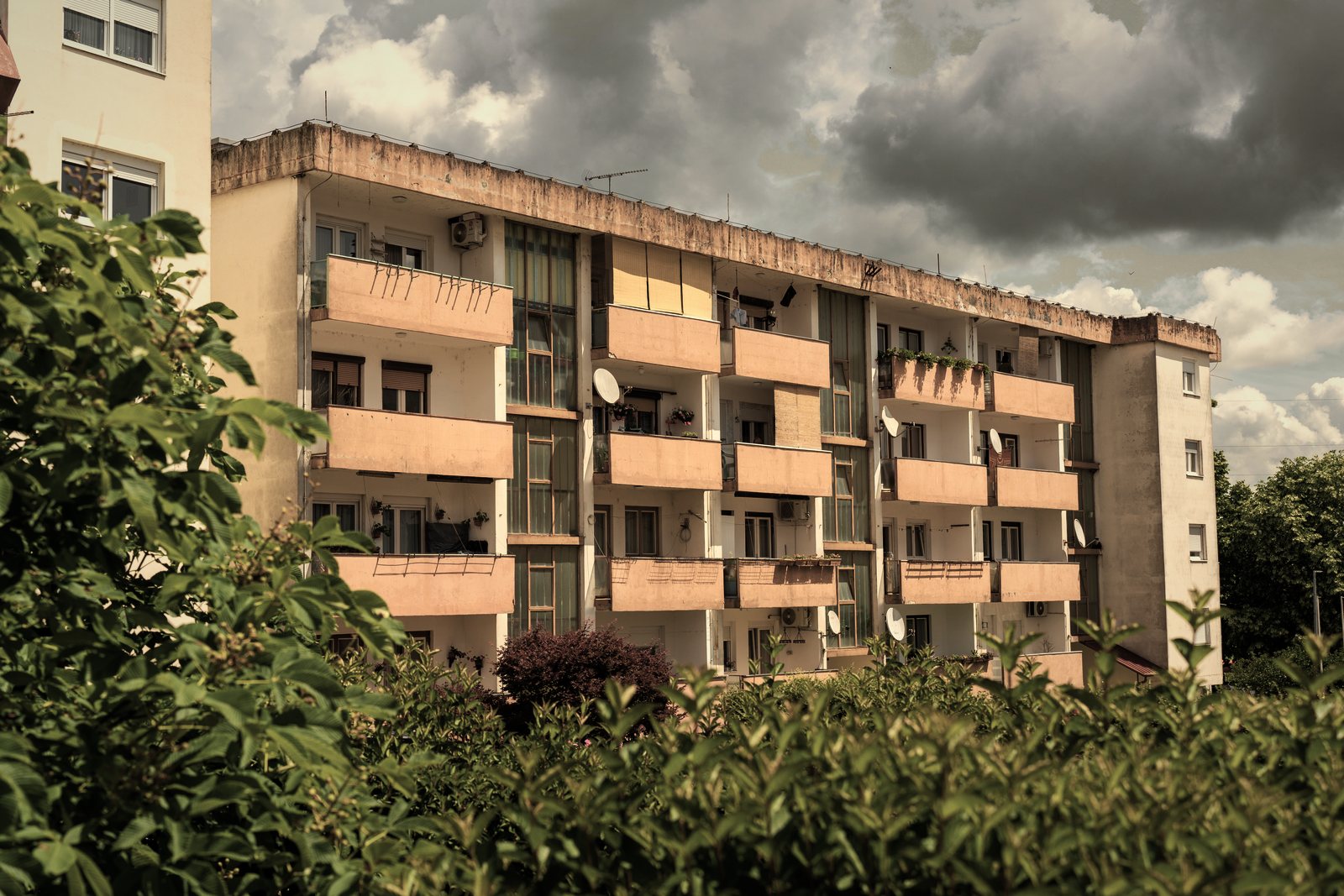 Immeuble collectif des années 1960 à balcons saumon, derrière une haie verte sous un ciel nuageux, photo d'illustration