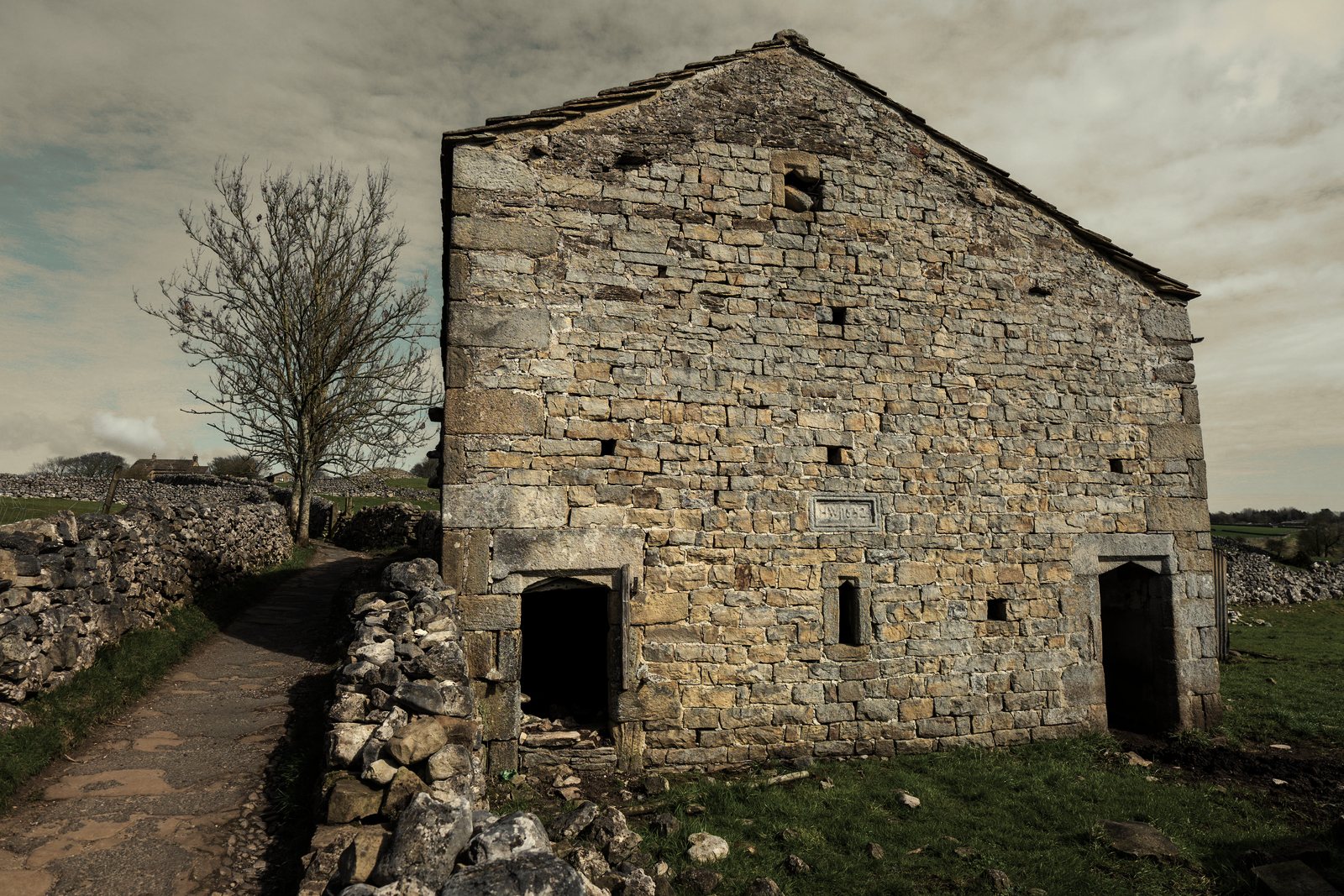 Grand bâtiment ancien en moellons de pierre calcaire à deux portes, le long d'un chemin bordé de murets, photo d'illustration