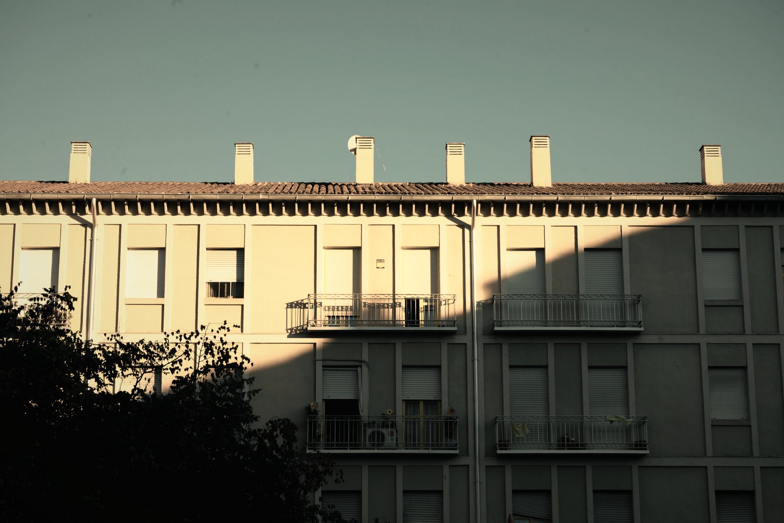 Façade d'immeuble des années 1950 à enduit clair, cheminées alignées et balcons, en contre-jour partiel, photo d'illustration