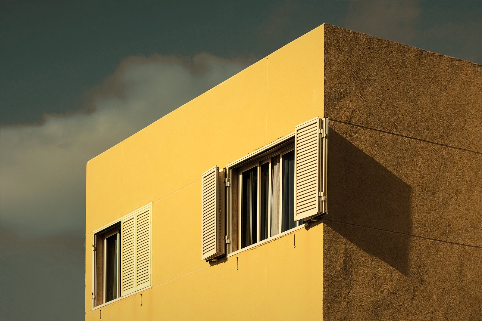 Angle d'un bâtiment récent à enduit jaune et volets persiennés blancs, sous un ciel nuageux, photo d'illustration